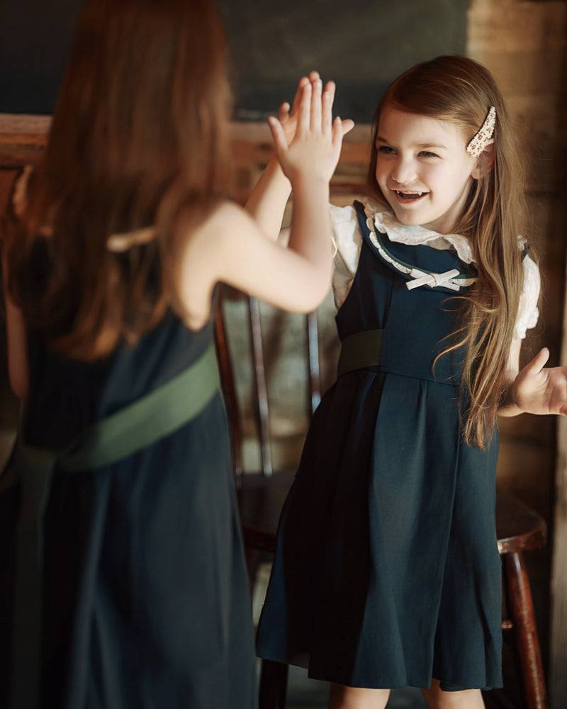 Two young girls in matching navy blue dresses with white collars and sashes pose for a high-five in a rustic indoor setting with warm, dim lighting.