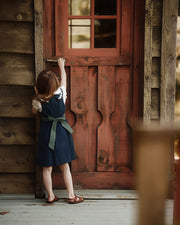 A young girl in a navy blue dress with a green sash stands on a rustic wooden porch, reaching toward a red-brown wooden door.
