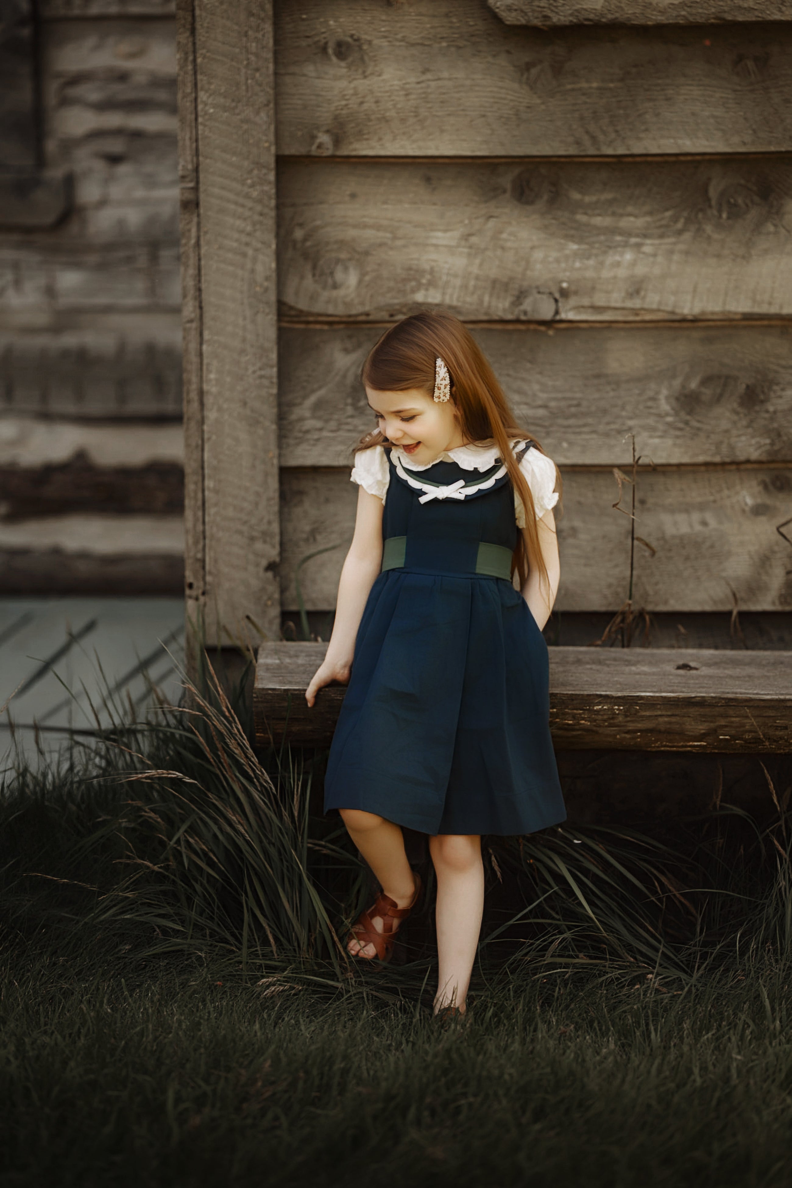 A young girl with long light brown hair stands outdoors in front of a rustic wooden log wall, wearing a navy blue pinafore-style dress with a white ruffled collar and sleeves, brown sandals, and a small hair clip.