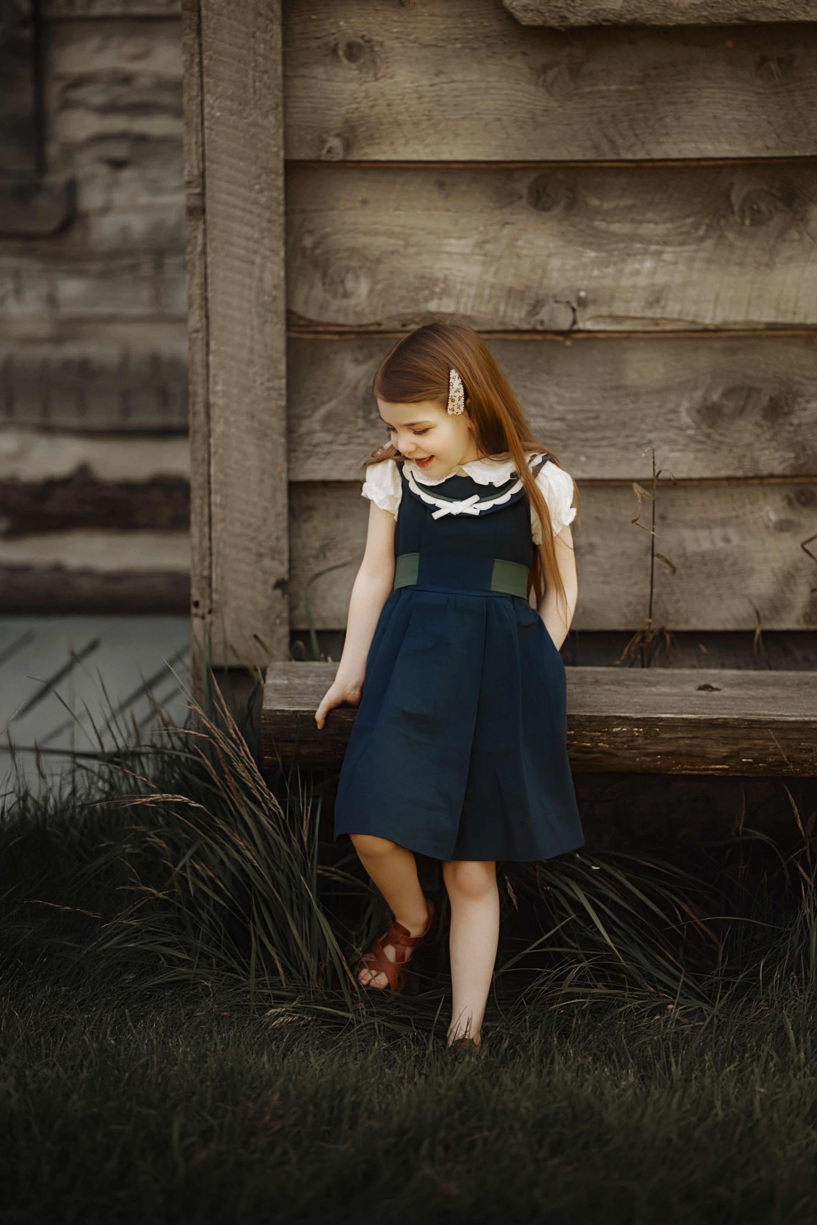 A young girl with long light brown hair stands outdoors in front of a rustic wooden log wall, wearing a navy blue pinafore-style dress with a white ruffled collar and sleeves, brown sandals, and a small hair clip.