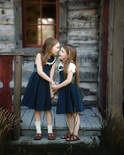 Two young girls in matching navy blue dresses with white collars and white short-sleeve tops stand on a rustic wooden porch in front of a weathered log cabin, holding hands and looking at each other.