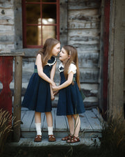 Two young girls in matching navy blue dresses with white collars and white short-sleeve tops stand on a rustic wooden porch in front of a weathered log cabin, holding hands and looking at each other.
