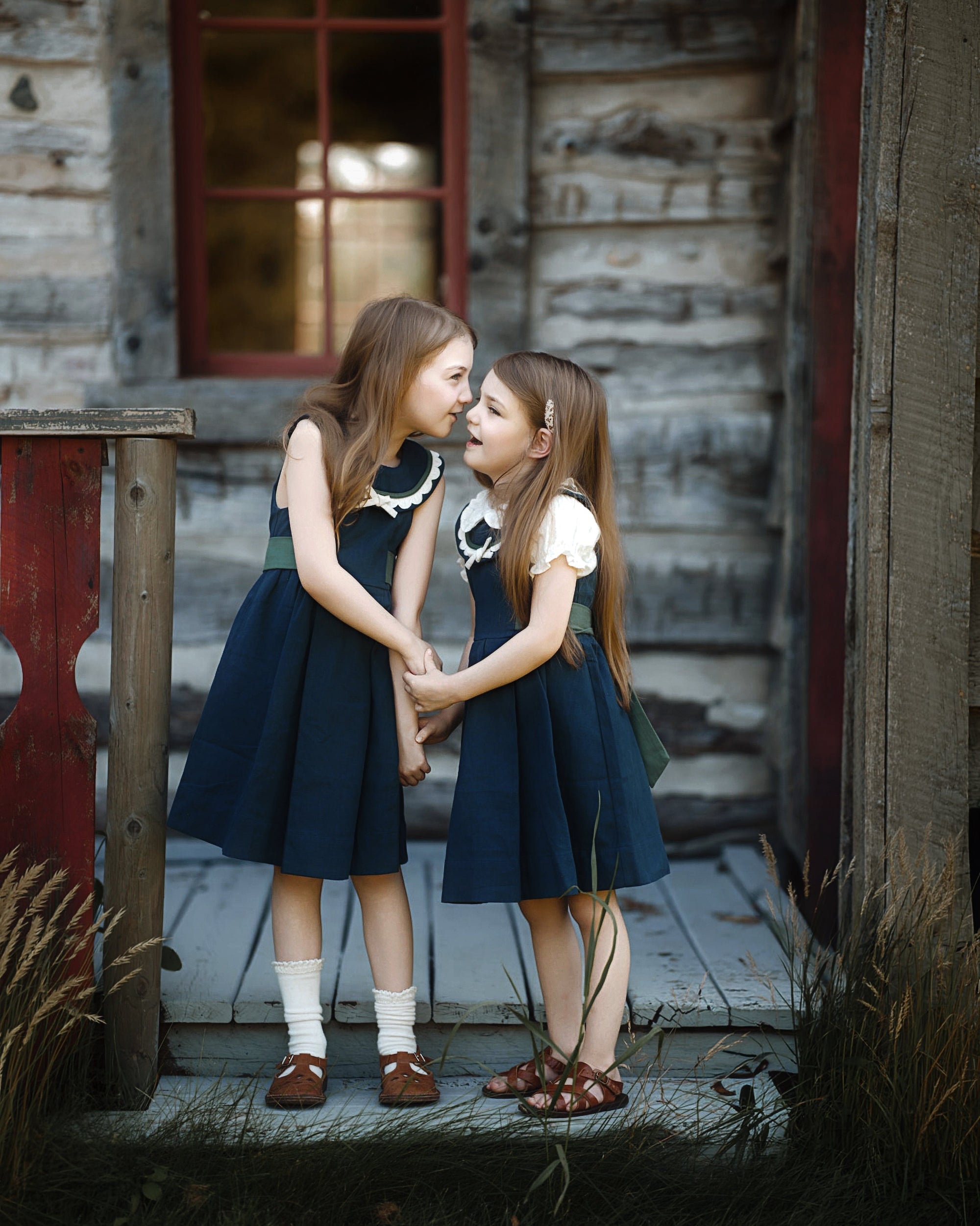Two young girls in matching navy blue dresses with white collars and white short-sleeve tops stand on a rustic wooden porch in front of a weathered log cabin, holding hands and looking at each other.