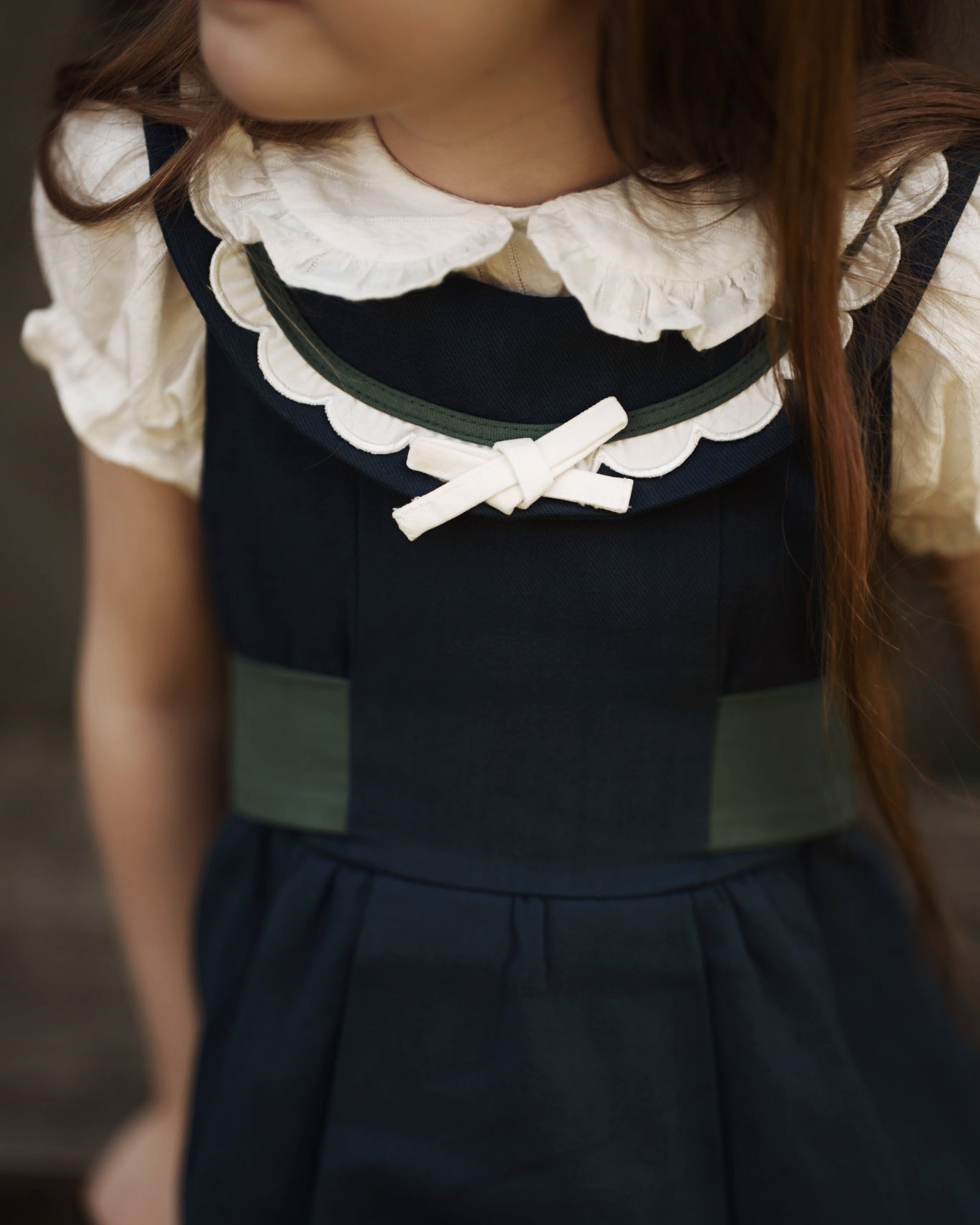 A close-up of a child wearing a navy blue dress with a white ruffled collar, a white bow at the neckline, and muted green side panels and trim.