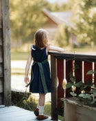 A young girl with light brown hair stands on a wooden porch, viewed from behind as she looks out over a garden. She is wearing a sleeveless navy blue dress with a dark green sash tied at the waist, white socks, and brown shoes.