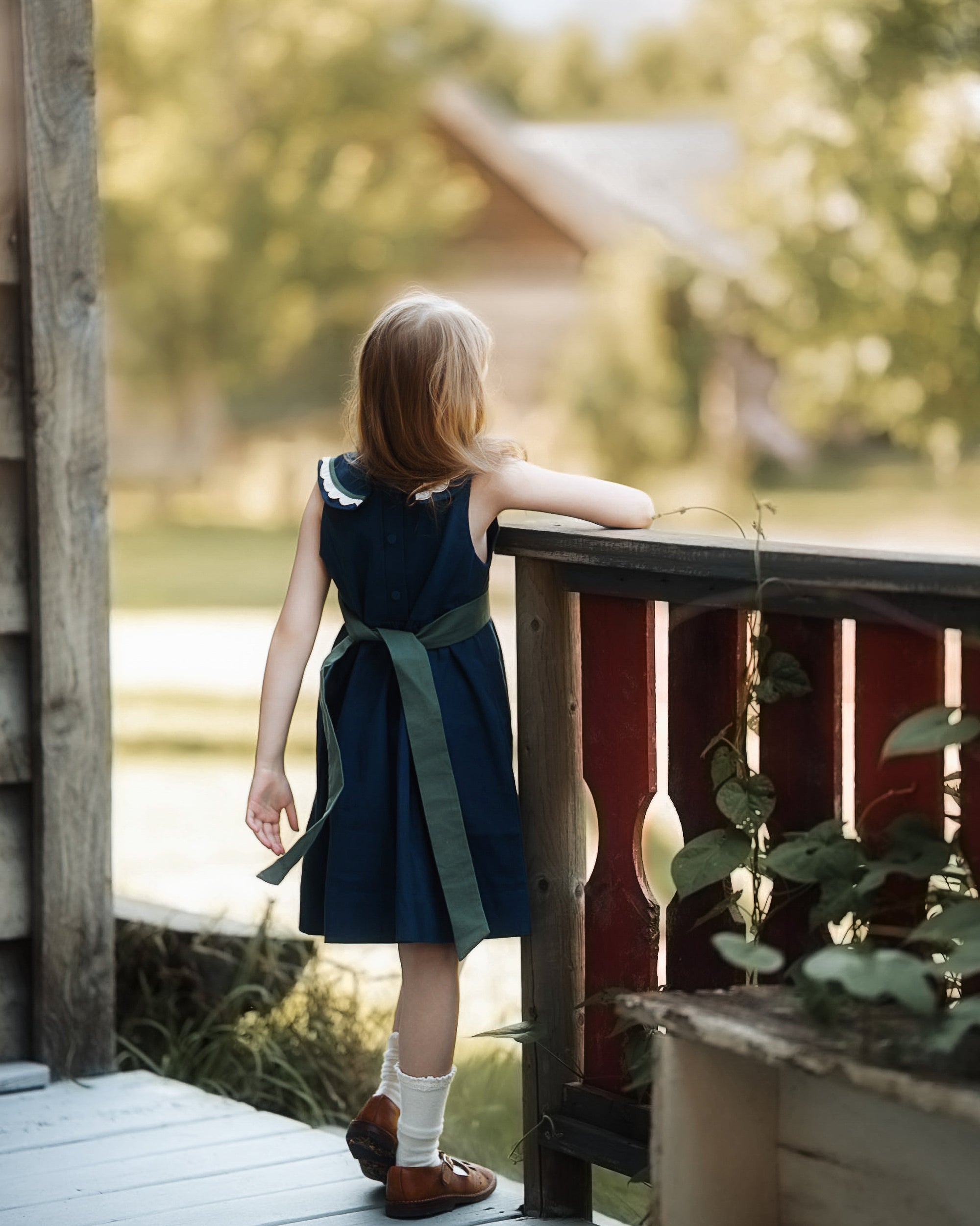 A young girl with light brown hair stands on a wooden porch, viewed from behind as she looks out over a garden. She is wearing a sleeveless navy blue dress with a dark green sash tied at the waist, white socks, and brown shoes.
