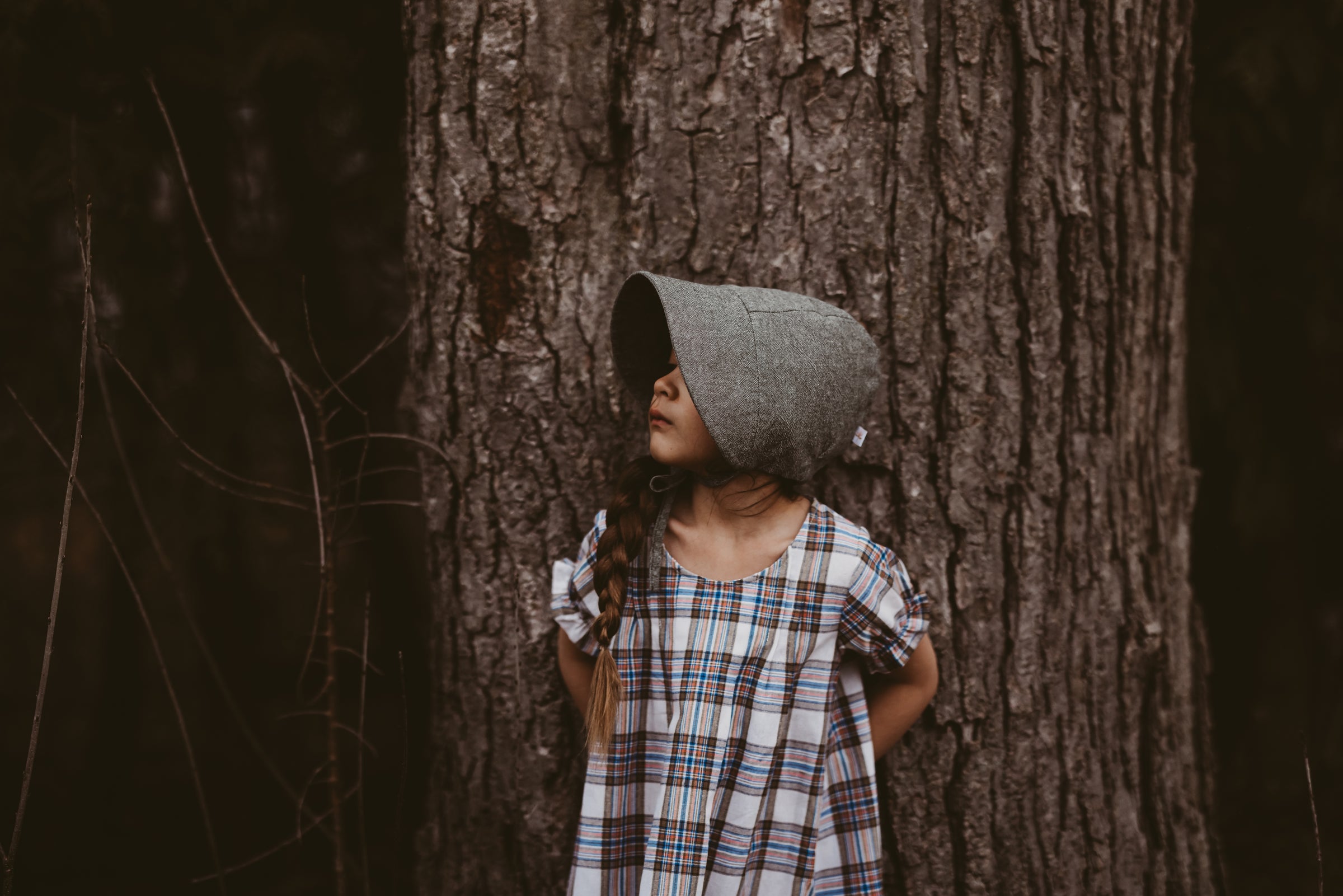 Girl in a plaid dress and green bonnet leaning against a tree