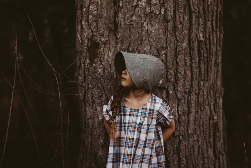 Girl in a plaid dress and green bonnet leaning against a tree