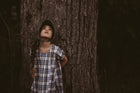 Girl in a plaid dress and green bonnet looking up leaning against a tree