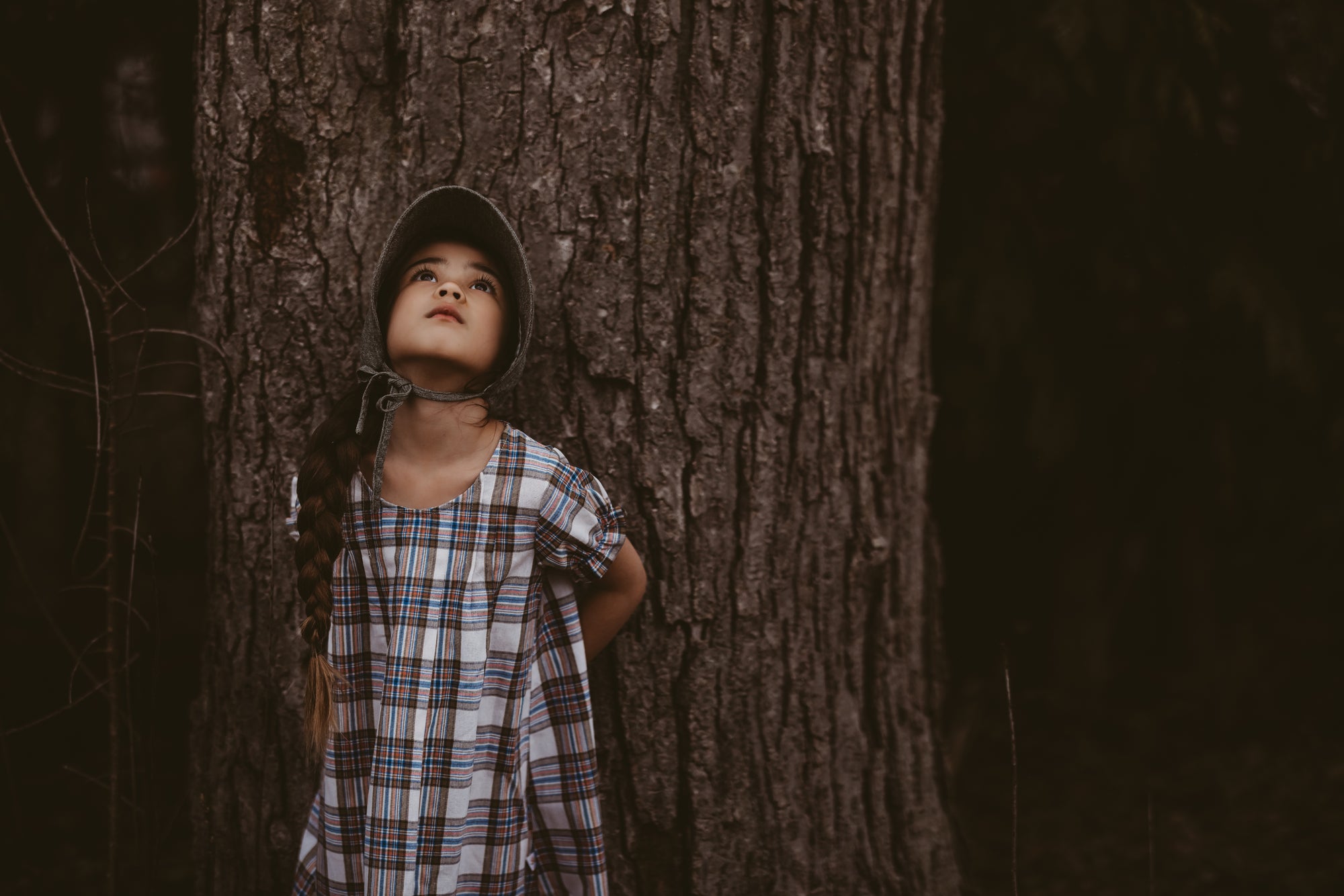 Girl in a plaid dress and green bonnet looking up leaning against a tree