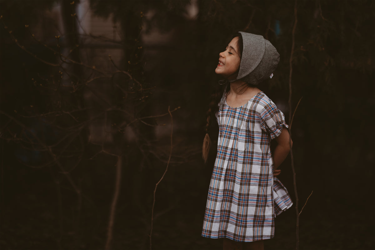 Girl wearing a plaid dress and a bonnet standing in a dark, forest-like setting.