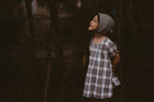 Girl wearing a plaid dress and a bonnet standing in a dark, forest-like setting.