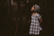 Girl wearing a plaid dress and a bonnet standing in a dark, forest-like setting.