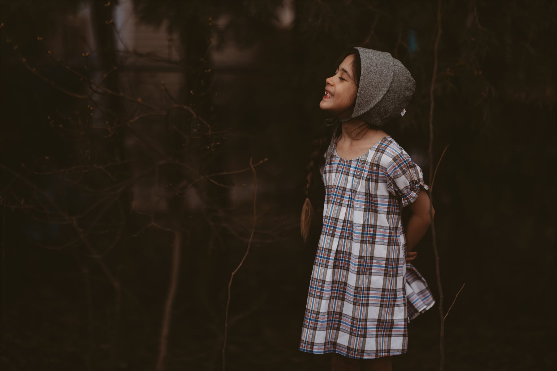 Girl wearing a plaid dress and a bonnet standing in a dark, forest-like setting.