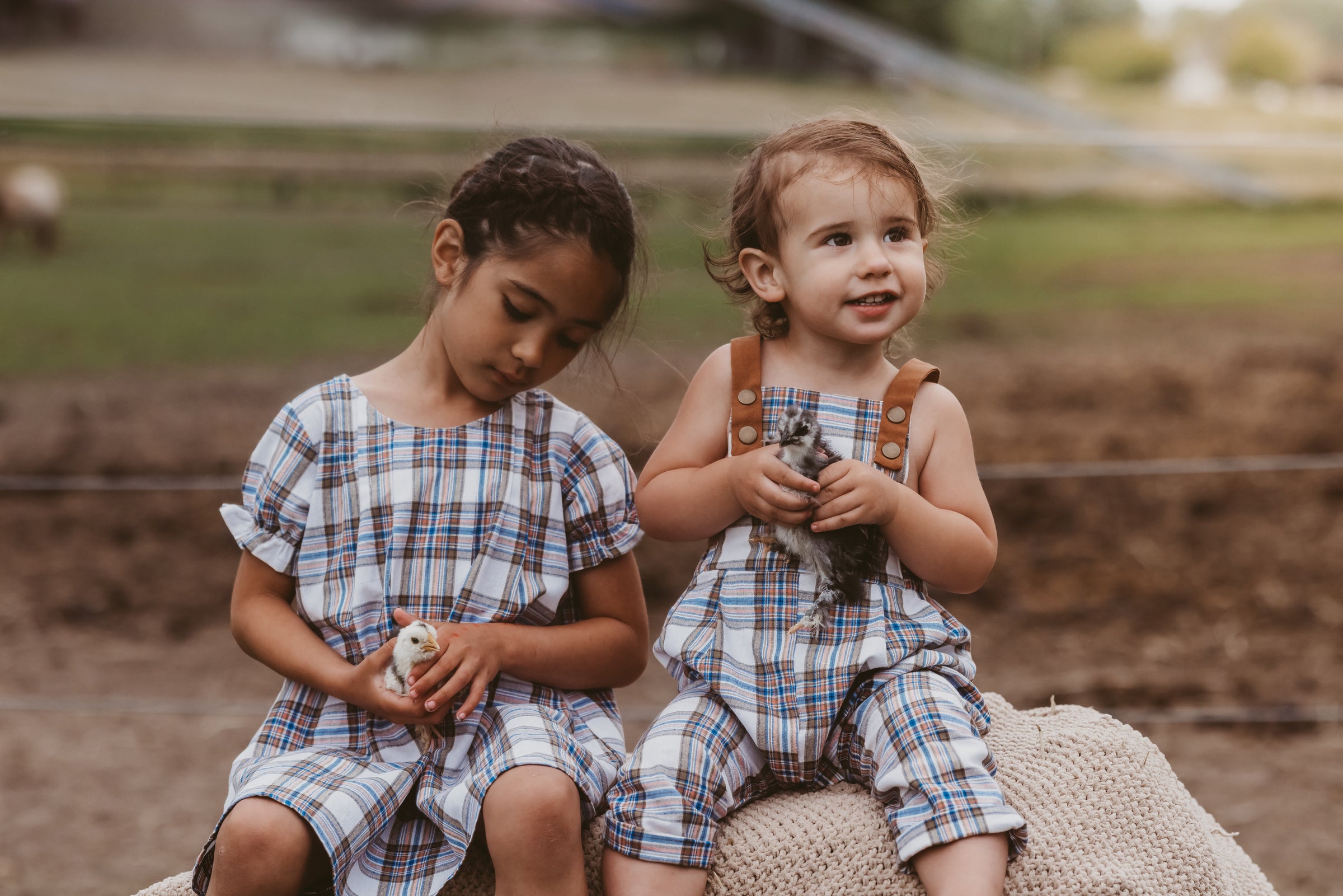 Two young girls in plaid outfits sitting on a rock outdoors.