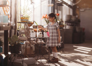 Young girl in a plaid dress standing in a sunlit room with plants and furniture.