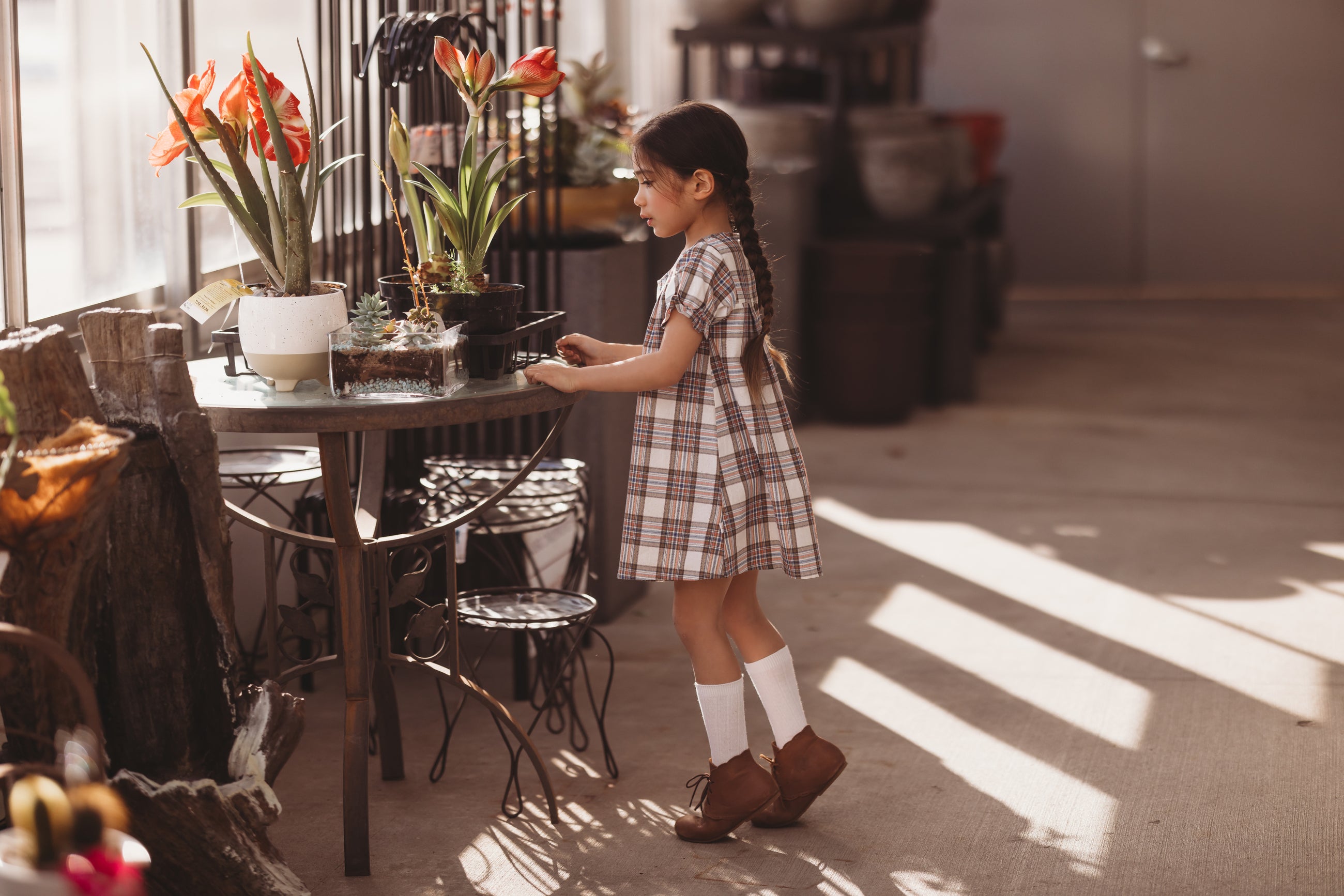 Young girl in a plaid dress standing near a table with flowers in a sunlit room.