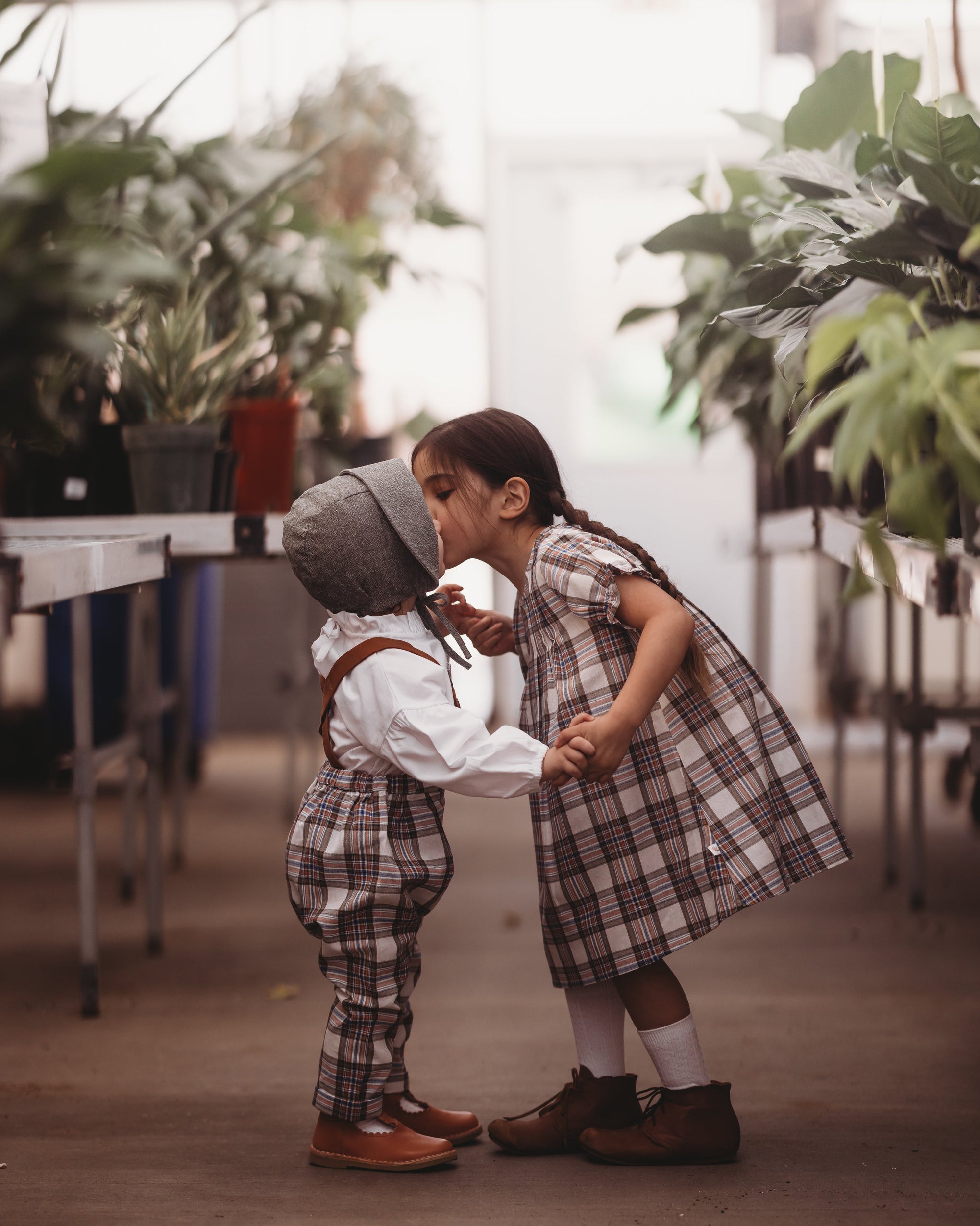 Two children in plaid outfits standing close together in a greenhouse.