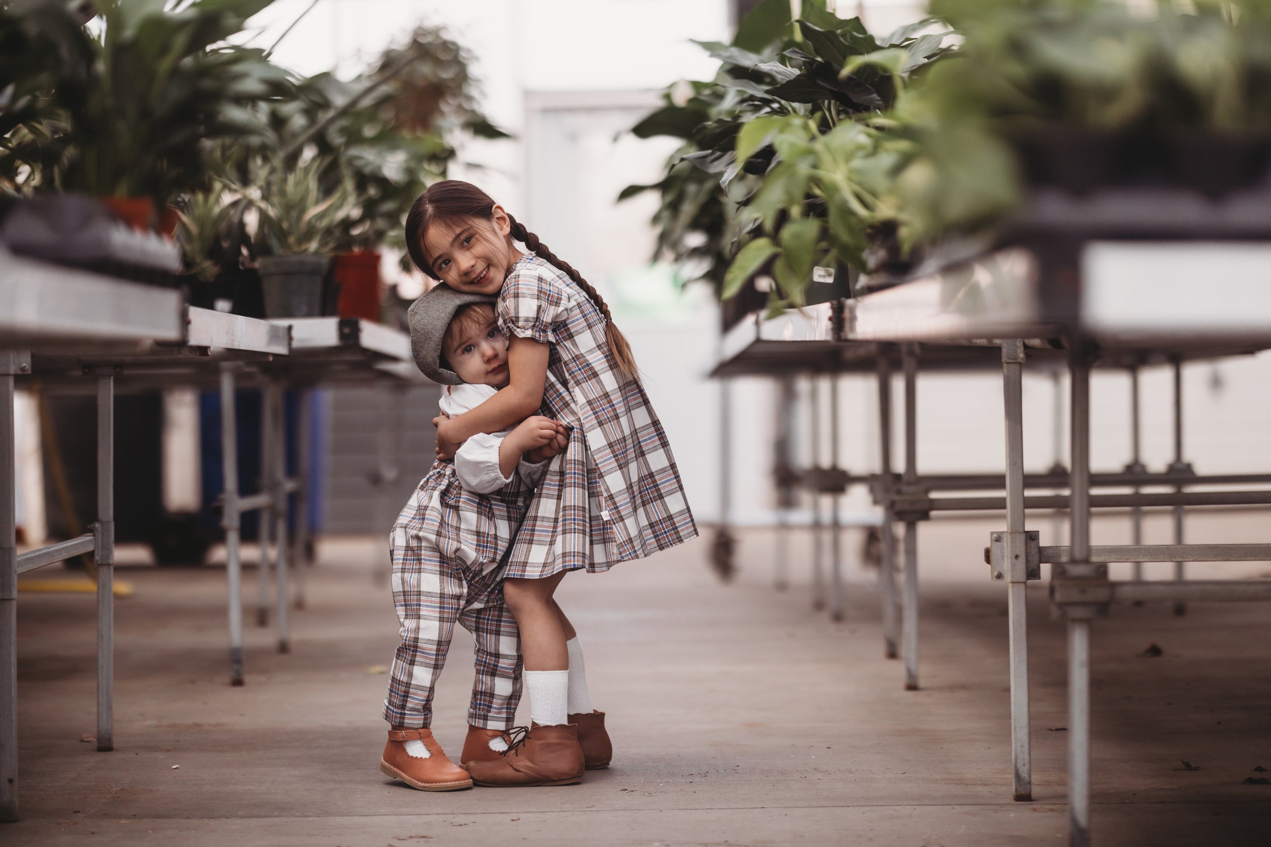 Two children in plaid outfits standing in a greenhouse.