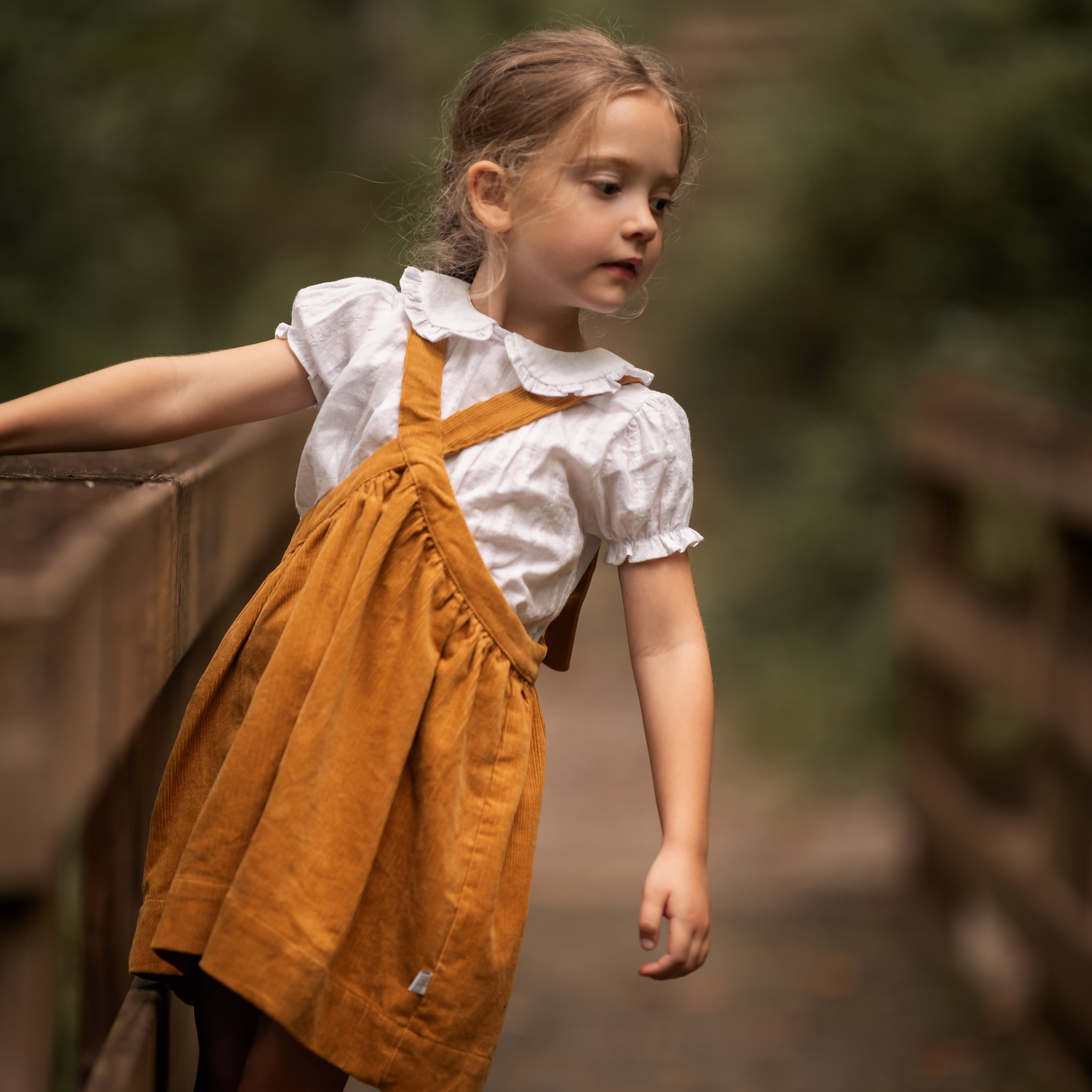Young girl in a yellow dress and white blouse standing on a wooden bridge with a blurred natural background