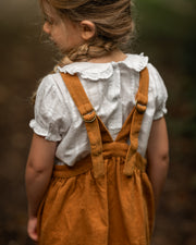 Child wearing a white blouse with a ruffled collar and brown overalls against a blurred natural background