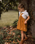 Young girl in a mustard pinafore dress standing next to a tree in a natural setting.