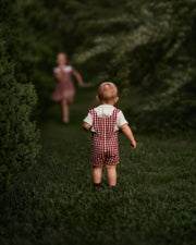 A toddler wearing a red and white gingham check overalls set with a white shirt walks away on a grassy path surrounded by dark green trees, with a blurred figure in a pink dress running in the background.