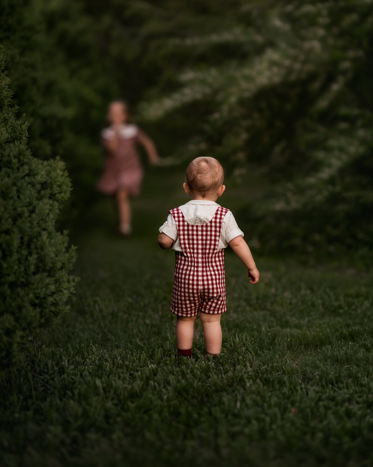 A toddler wearing a red and white gingham check overalls set with a white shirt walks away on a grassy path surrounded by dark green trees, with a blurred figure in a pink dress running in the background.