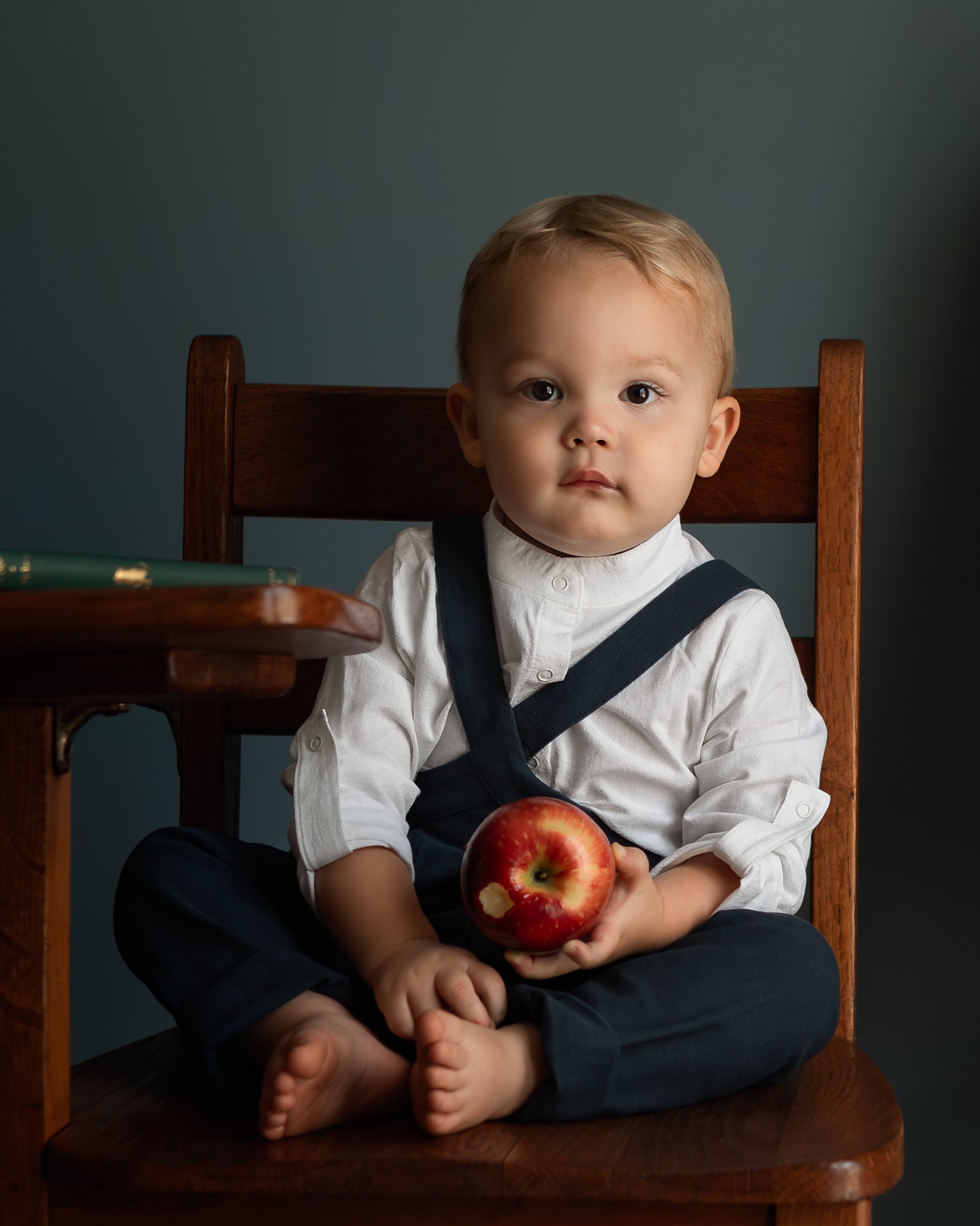 A baby boy sitting cross-legged on a wooden chair, wearing a white long-sleeve shirt and dark navy overalls while holding a red apple.