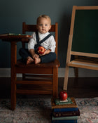 A young child sits cross-legged on a wooden school desk chair holding an apple, with a dark chalkboard easel and books on a patterned rug in the background.