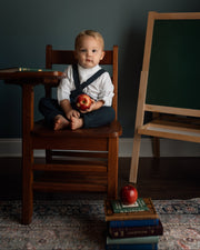 A young child sits cross-legged on a wooden school desk chair holding an apple, with a dark chalkboard easel and books on a patterned rug in the background.