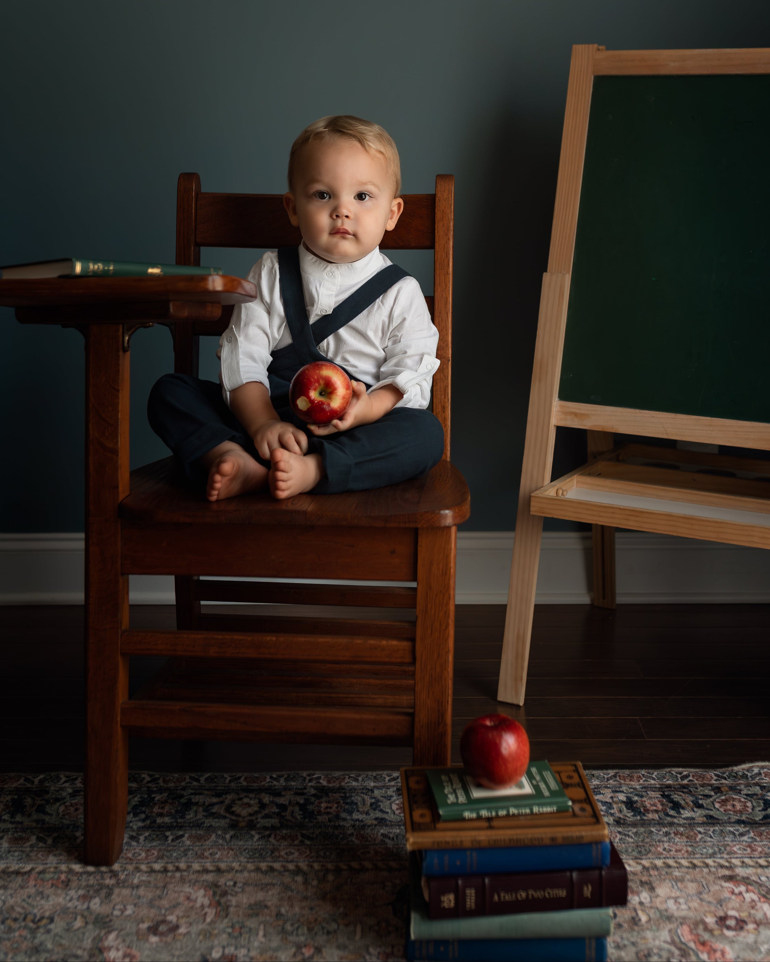A young child sits cross-legged on a wooden school desk chair holding an apple, with a dark chalkboard easel and books on a patterned rug in the background.