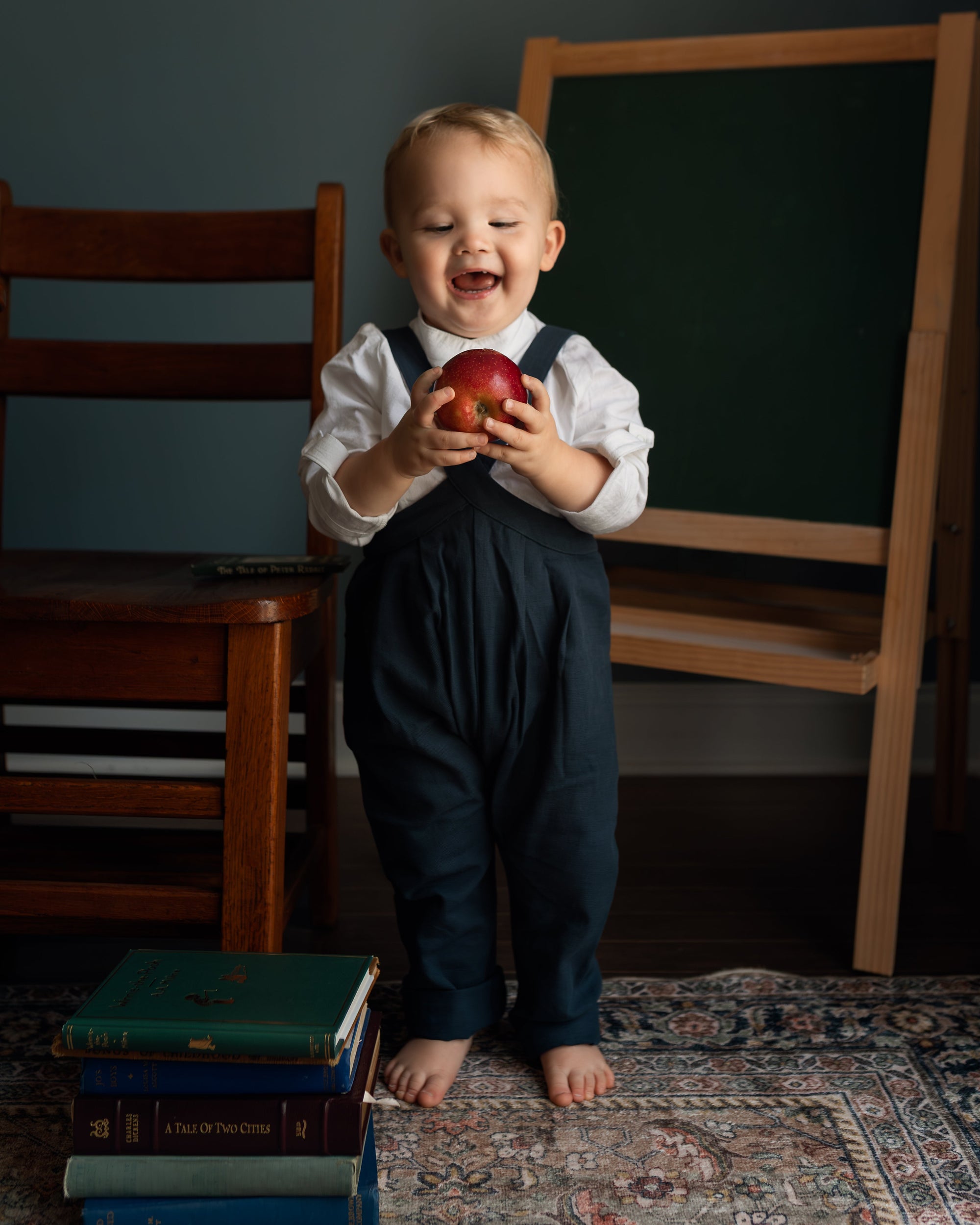 A toddler stands barefoot on a patterned rug in a room, wearing a white long-sleeve shirt and dark navy overalls, holding a red apple. A wooden chair, a stack of books, and a chalkboard on a stand are visible in the background.