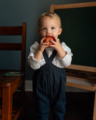 A toddler boy wearing a white long-sleeve shirt and navy blue overalls stands in front of a dark chalkboard, holding and biting into a red apple.