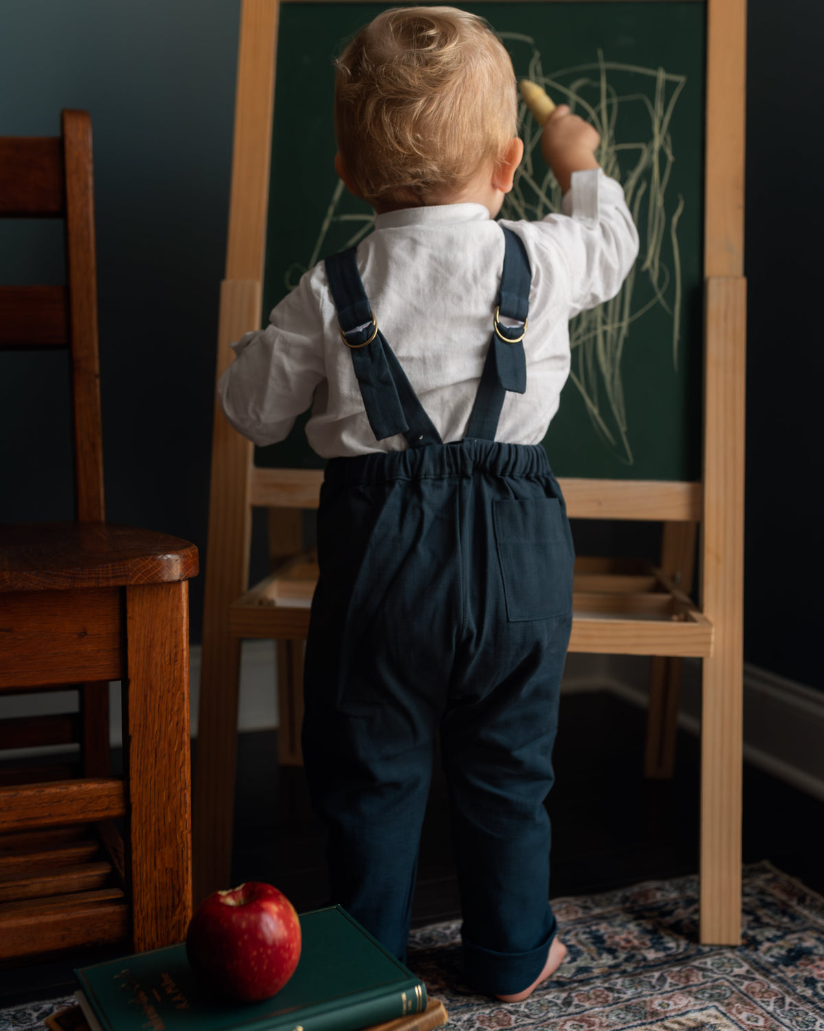 A toddler standing with their back to the camera, wearing a white long-sleeve shirt and navy blue overalls, drawing on a wooden easel chalkboard. The scene is set in a room with a dark teal wall, a wooden chair, a patterned rug, and a stack of books with a red apple on top.