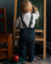 A toddler standing with their back to the camera, wearing a white long-sleeve shirt and navy blue overalls, drawing on a wooden easel chalkboard. The scene is set in a room with a dark teal wall, a wooden chair, a patterned rug, and a stack of books with a red apple on top.