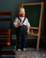 A toddler stands barefoot in a room with a dark wall, holding a red apple to their mouth. The child is wearing a white long-sleeve shirt and navy blue overalls.