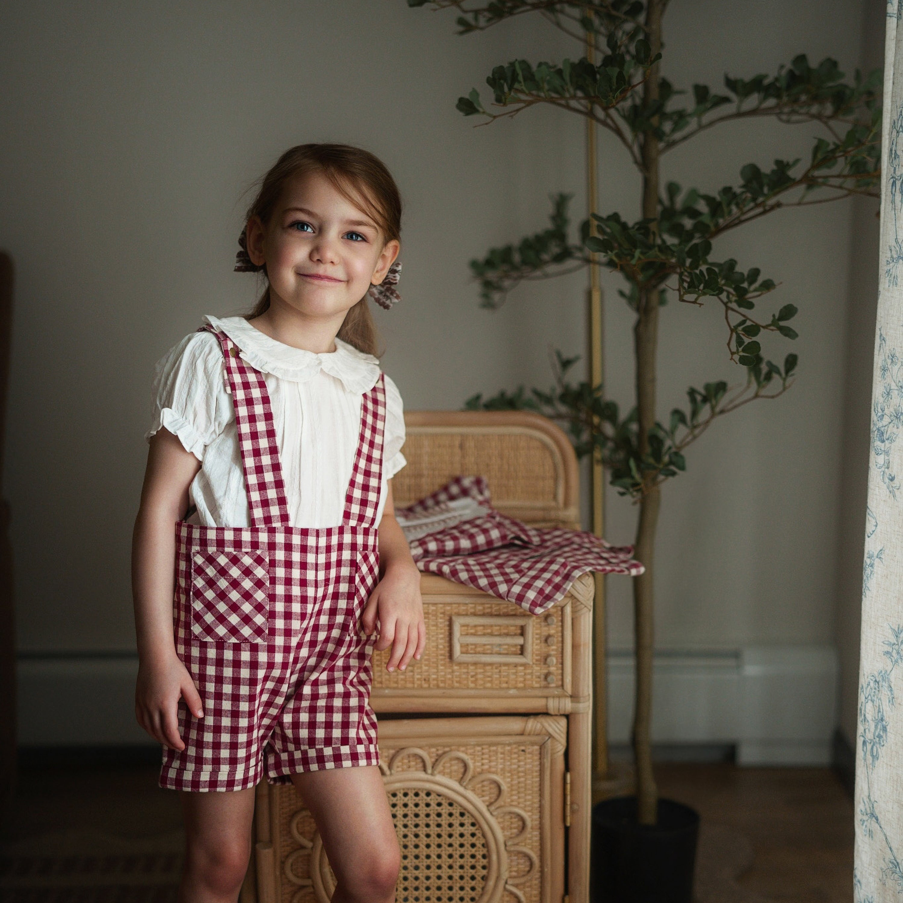 Young girl in a red checkered dress standing in a room with a wicker chair and plant.