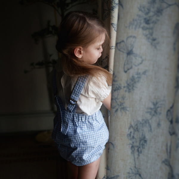 Child in blue checkered overalls standing near a floral curtain