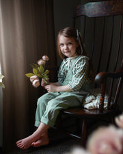 Young girl in a green dress sitting on a wooden chair holding flowers, with a blurred foreground.