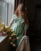 Woman in a green dress and apron sitting by a window with flowers