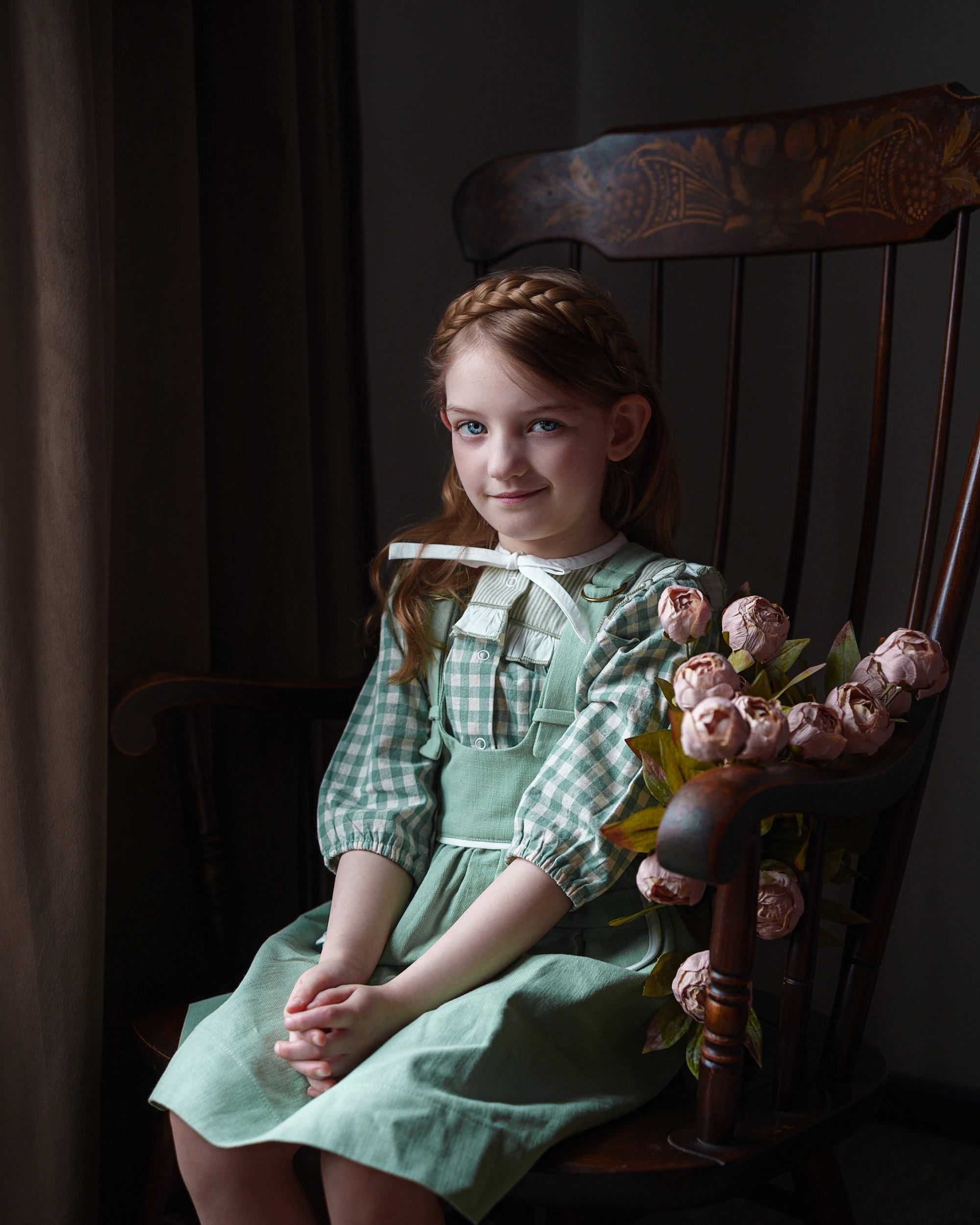 Young girl in a green dress sitting on a wooden chair with flowers, in a dimly lit room.
