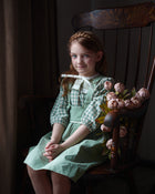 Young girl in a green dress sitting on a wooden chair with flowers, in a dimly lit room.