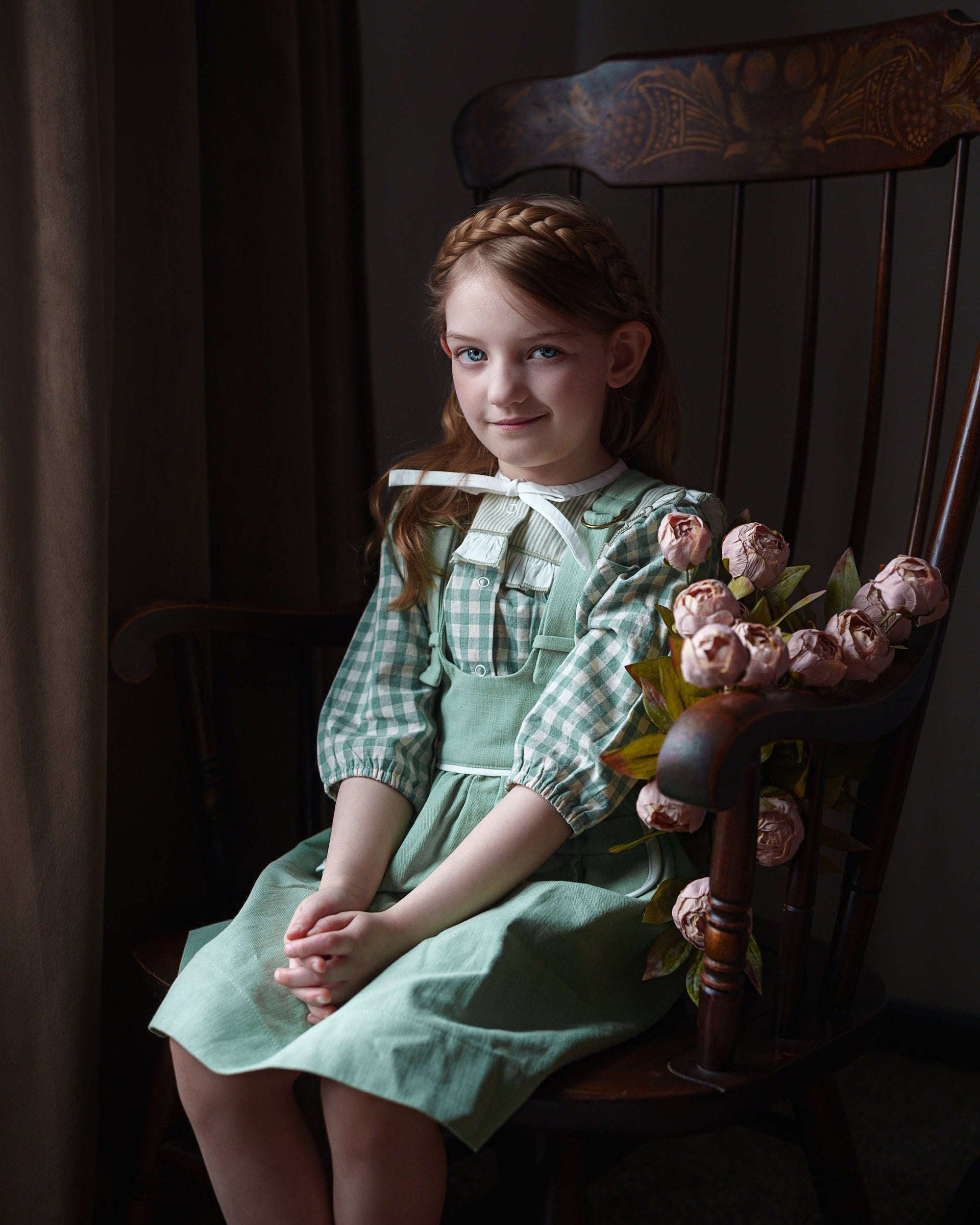 Young girl in a green dress sitting on a wooden chair with flowers, in a dimly lit room.