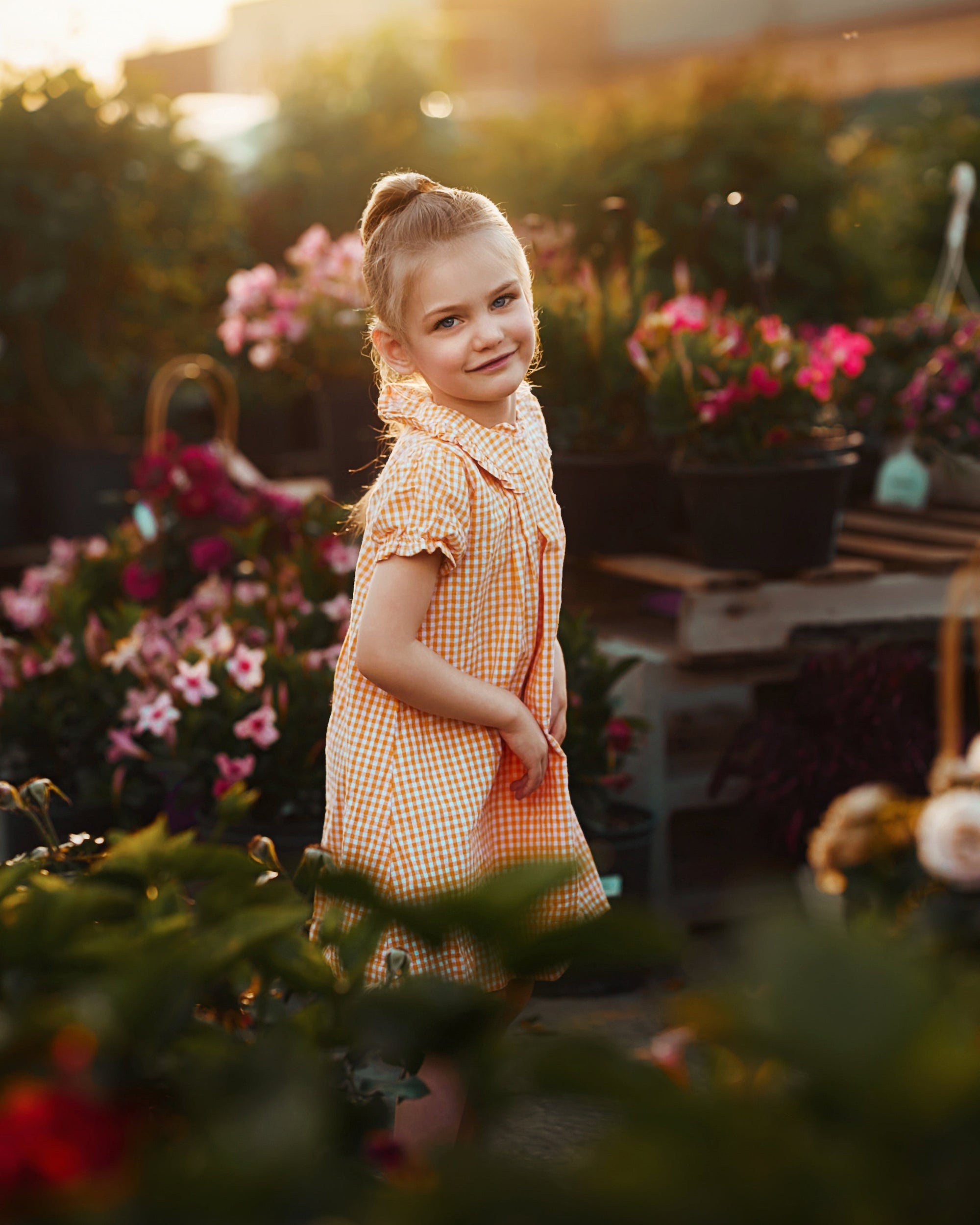 A young girl with blonde hair in a ponytail stands in a flower garden at sunset, wearing a yellow and white gingham dress. The background is filled with colorful flowers and green foliage, with warm golden sunlight filtering through.