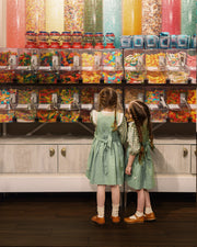 Two young girls in matching sage green pinafore dresses stand in front of a large candy display, looking up at tall transparent bins filled with colorful sprinkles and gummy candies in a candy store.