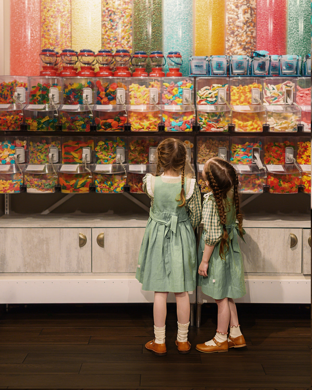 Two young girls in matching sage green pinafore dresses stand in front of a large candy display, looking up at tall transparent bins filled with colorful sprinkles and gummy candies in a candy store.