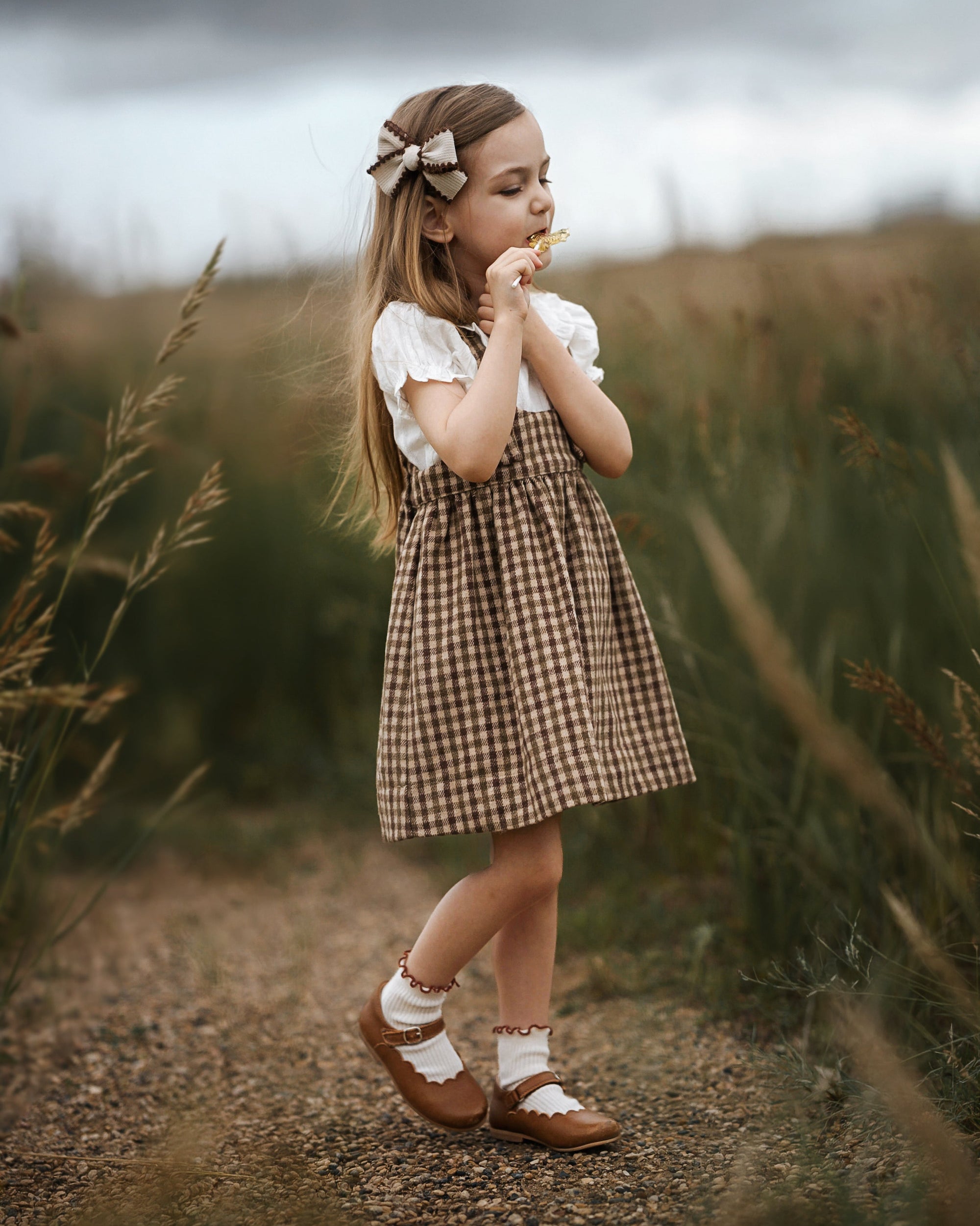 A young girl stands on a dirt path in a grassy field, wearing a brown and beige gingham pinafore dress over a white short-sleeve top, with brown Mary Jane shoes and a beige bow in her hair.