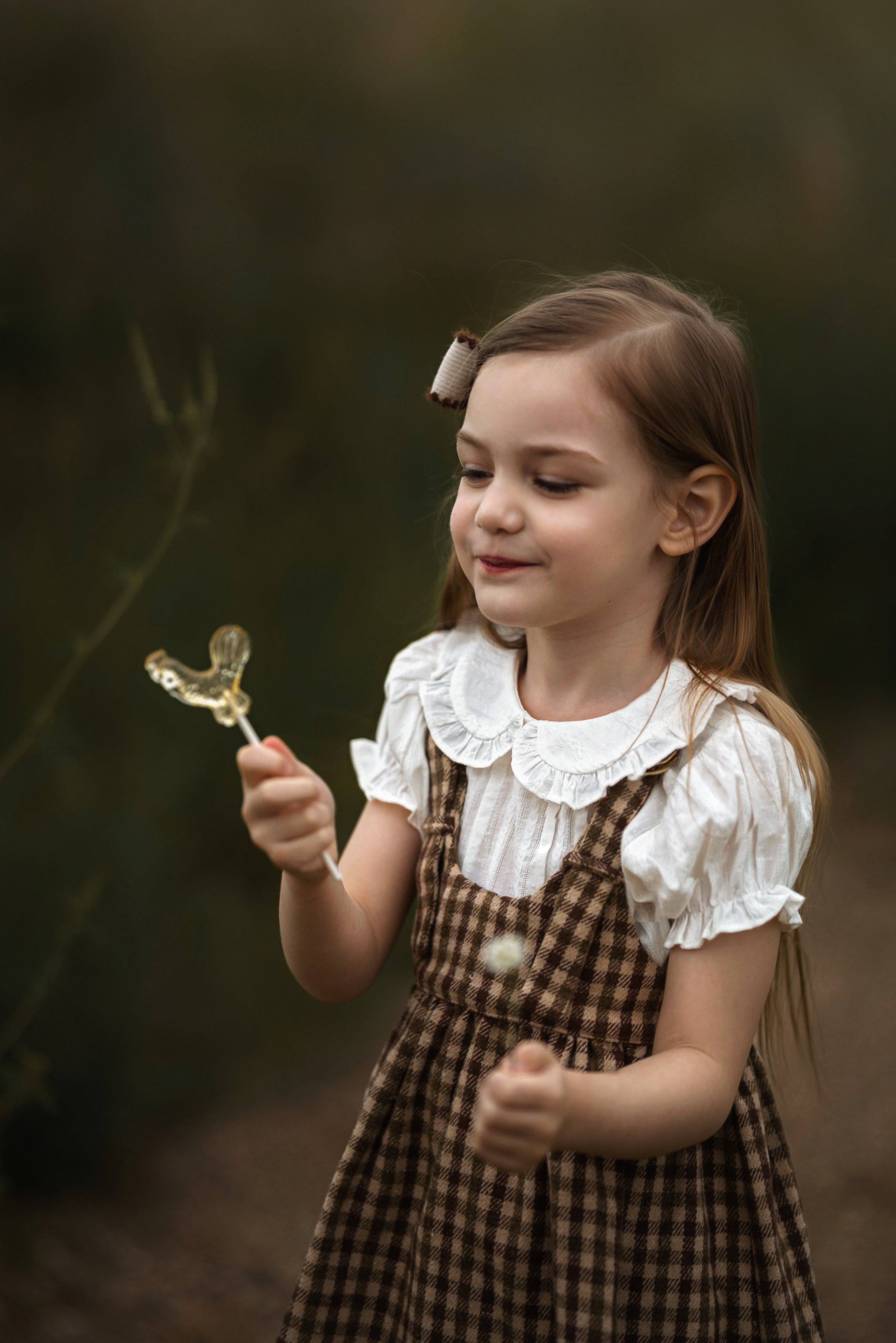 A young girl with long brown hair stands outdoors in a forest-like setting, wearing a white ruffled blouse and a brown plaid pinafore dress. She is holding a small gold butterfly-shaped object in one hand and a dandelion in the other.