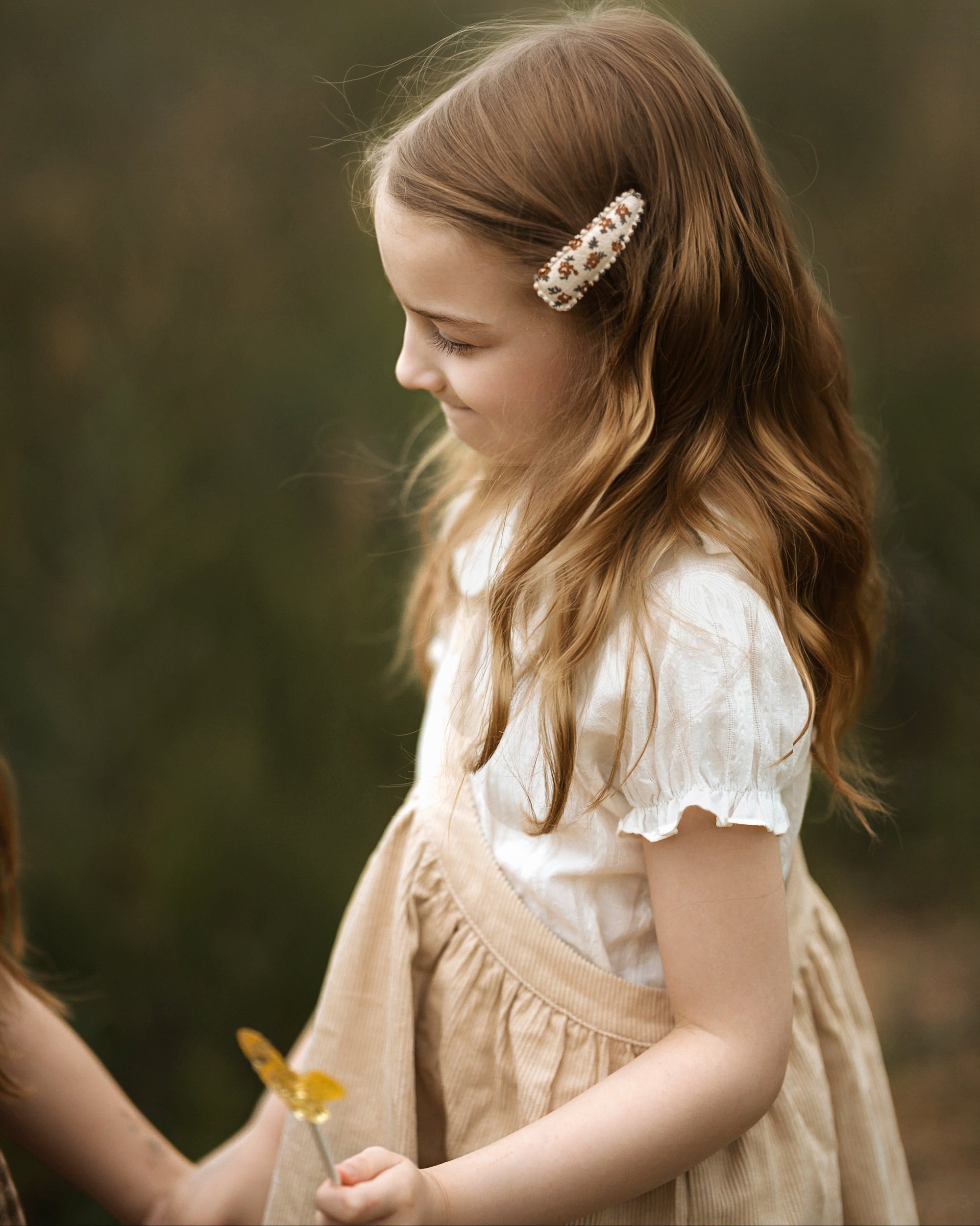 A young girl with long light brown hair wearing a white ruffled blouse and beige pinafore dress, holding a yellow flower. She is wearing a decorative pearl and gold barrette hair clip.